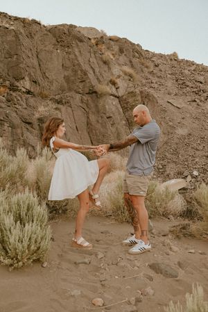 A couple dances playfully against a desert cliff backdrop, with the woman in a white dress and man in grey casual wear.