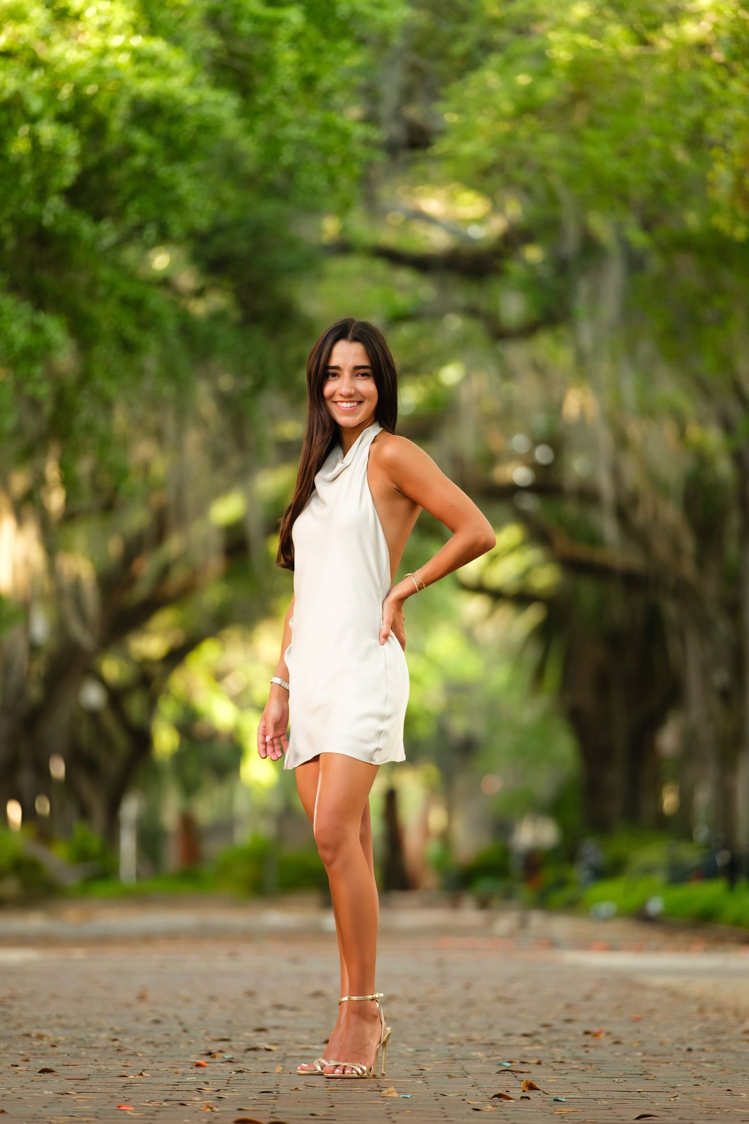 UF grad in white poses with her hand on her hip beneath a tree lined canopy.