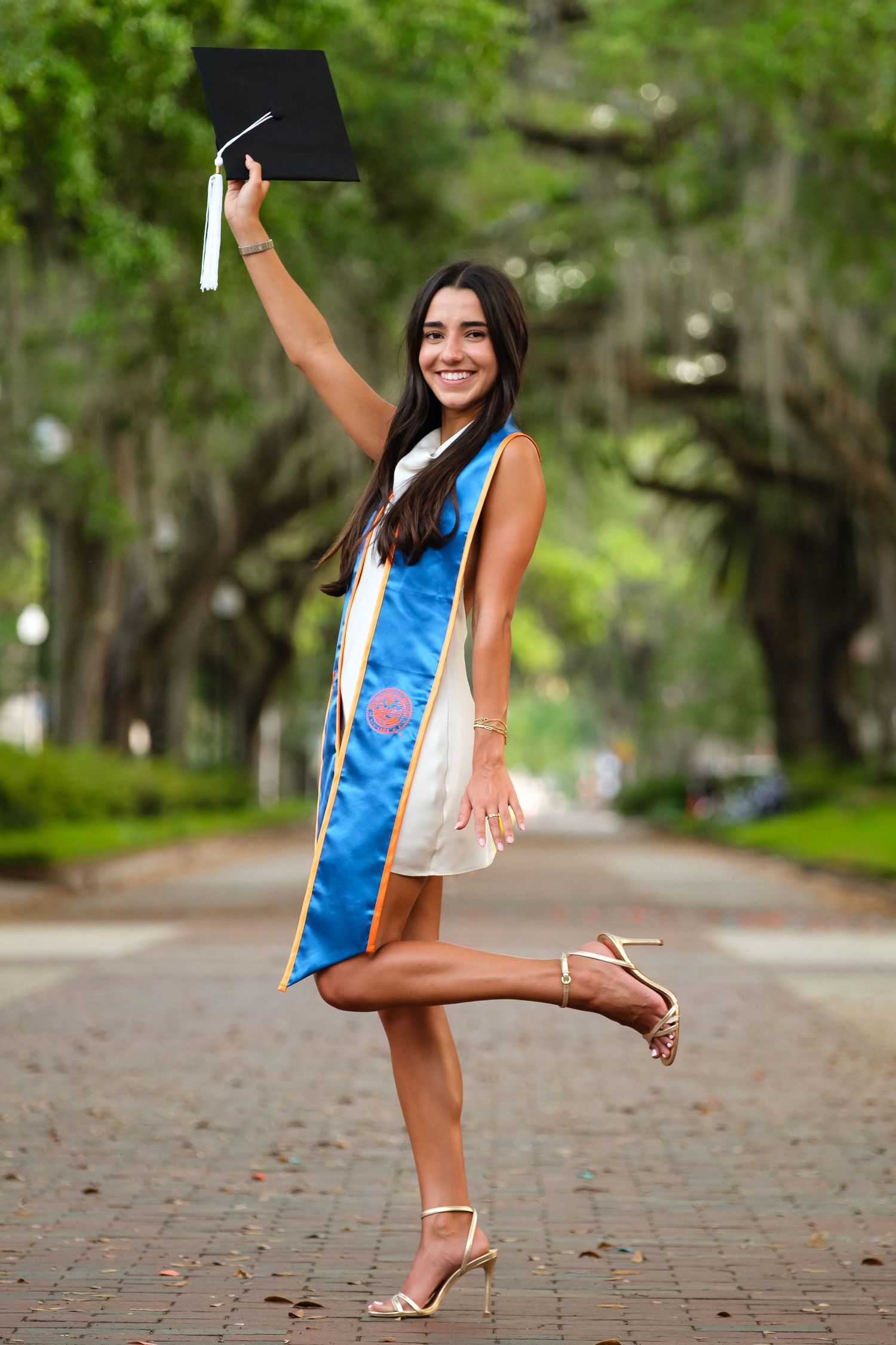 A smiling UF grad poses holding her cap and wearing a blue stole standing on one leg on a tree lined walkway at UF.