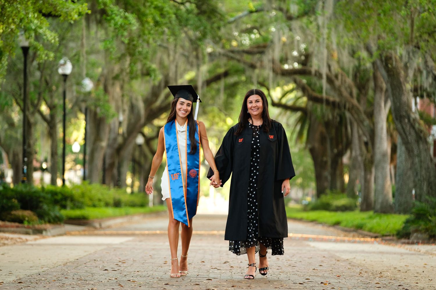 A smiling UF grad walks towards the camera holding hands with her mother who is wearing her gown.
