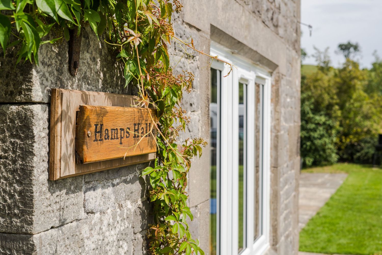A rustic wooden sign reading 'Hauses Edge' mounted on a stone wall with climbing vines and white window frame.
