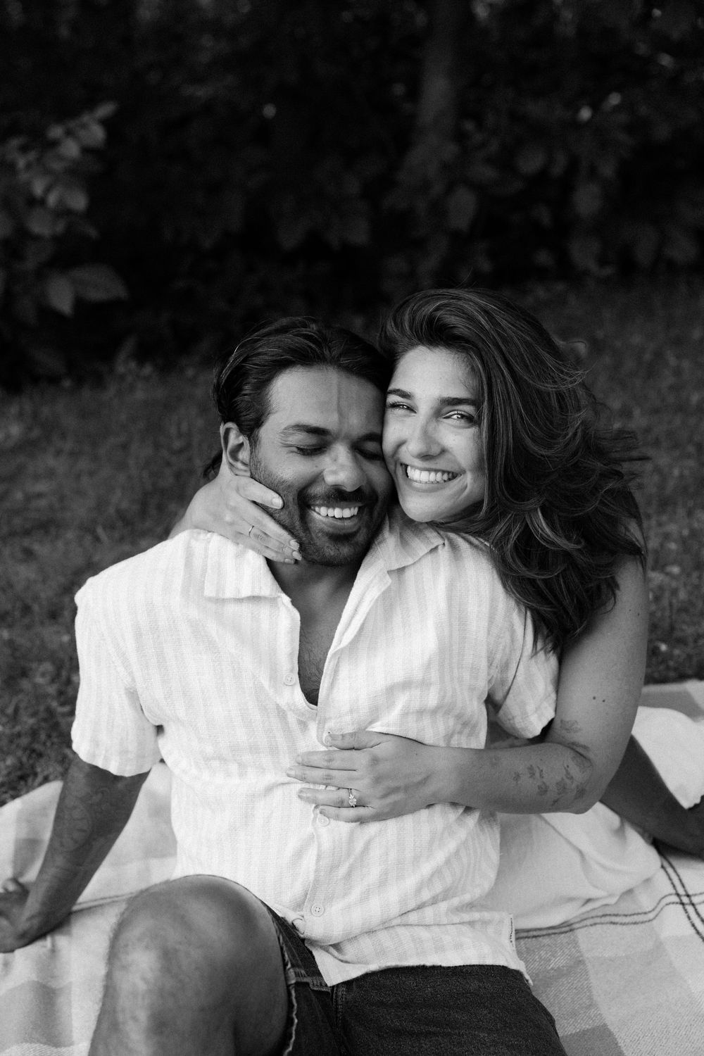 Black and white photo of a joyful couple sharing laughter while sitting on a blanket outdoors.