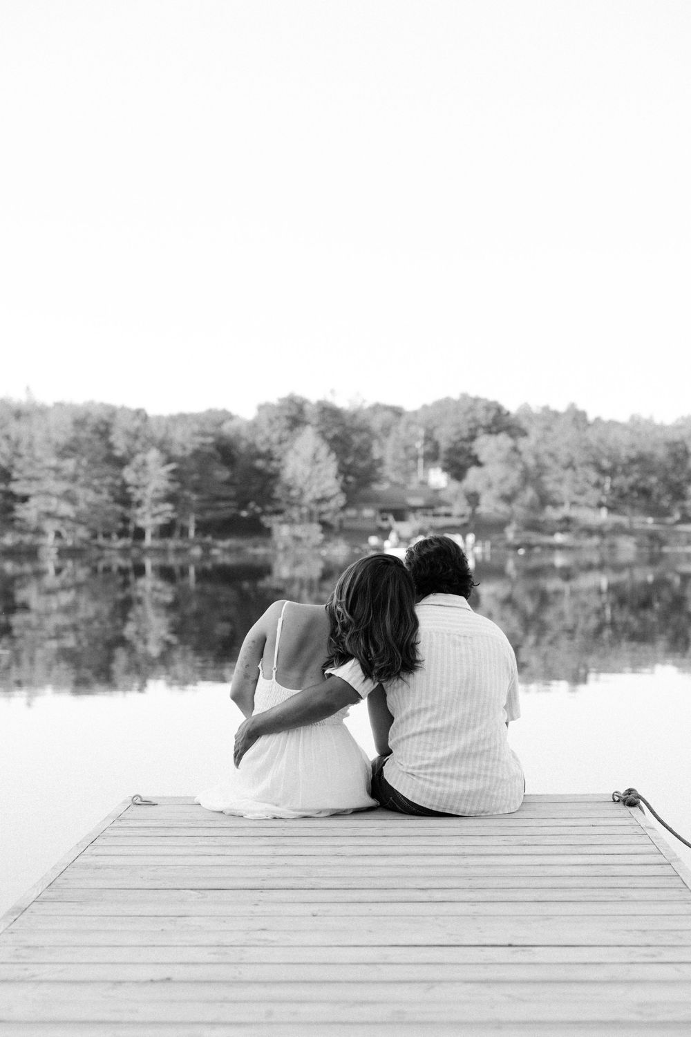 A couple lounges peacefully on a wooden dock overlooking a serene lake on a calm summer day.