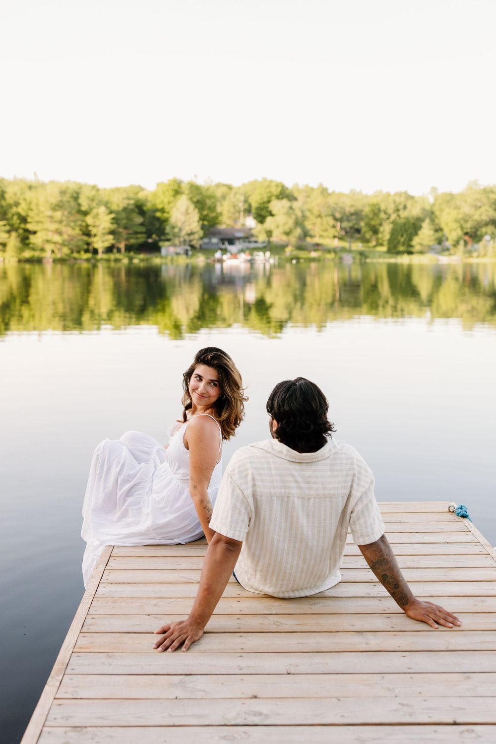 A couple in white sits together on a wooden dock by a serene lake surrounded by trees on a summer day.