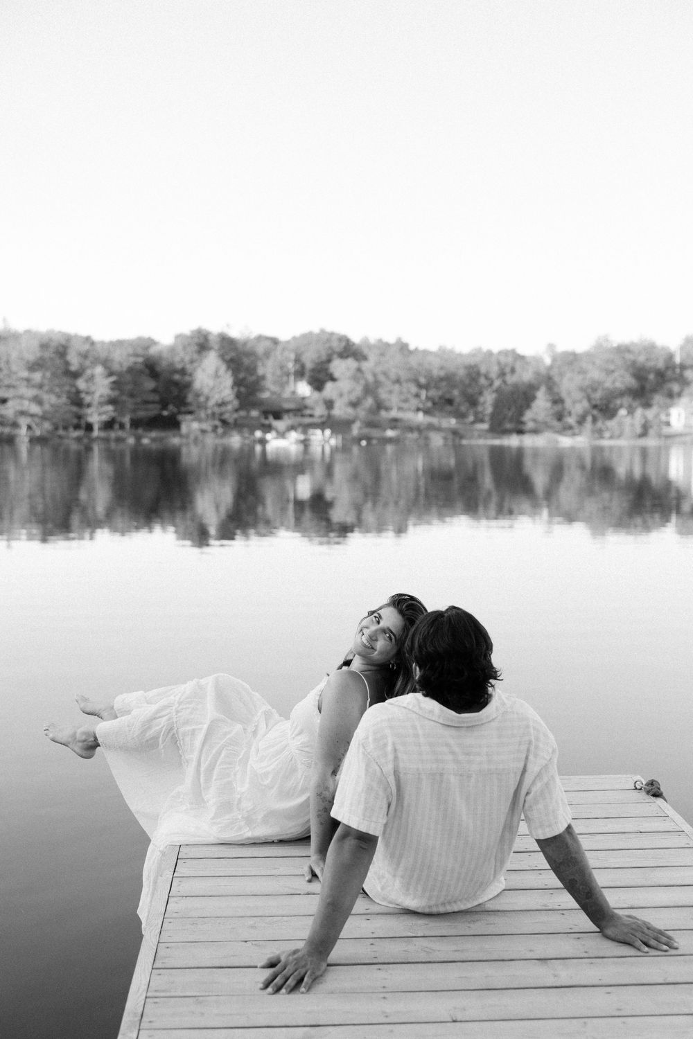 A romantic couple sits together on a wooden dock overlooking a serene lake with trees in the background.