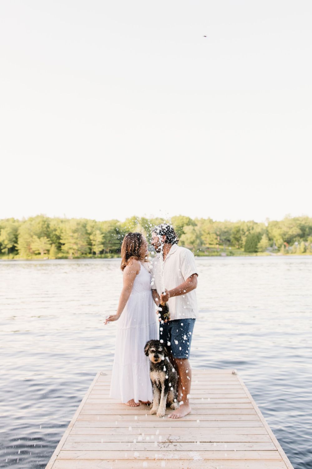 A couple shares a romantic moment on a wooden dock by the lake as flower petals fall around them in the sunlight.