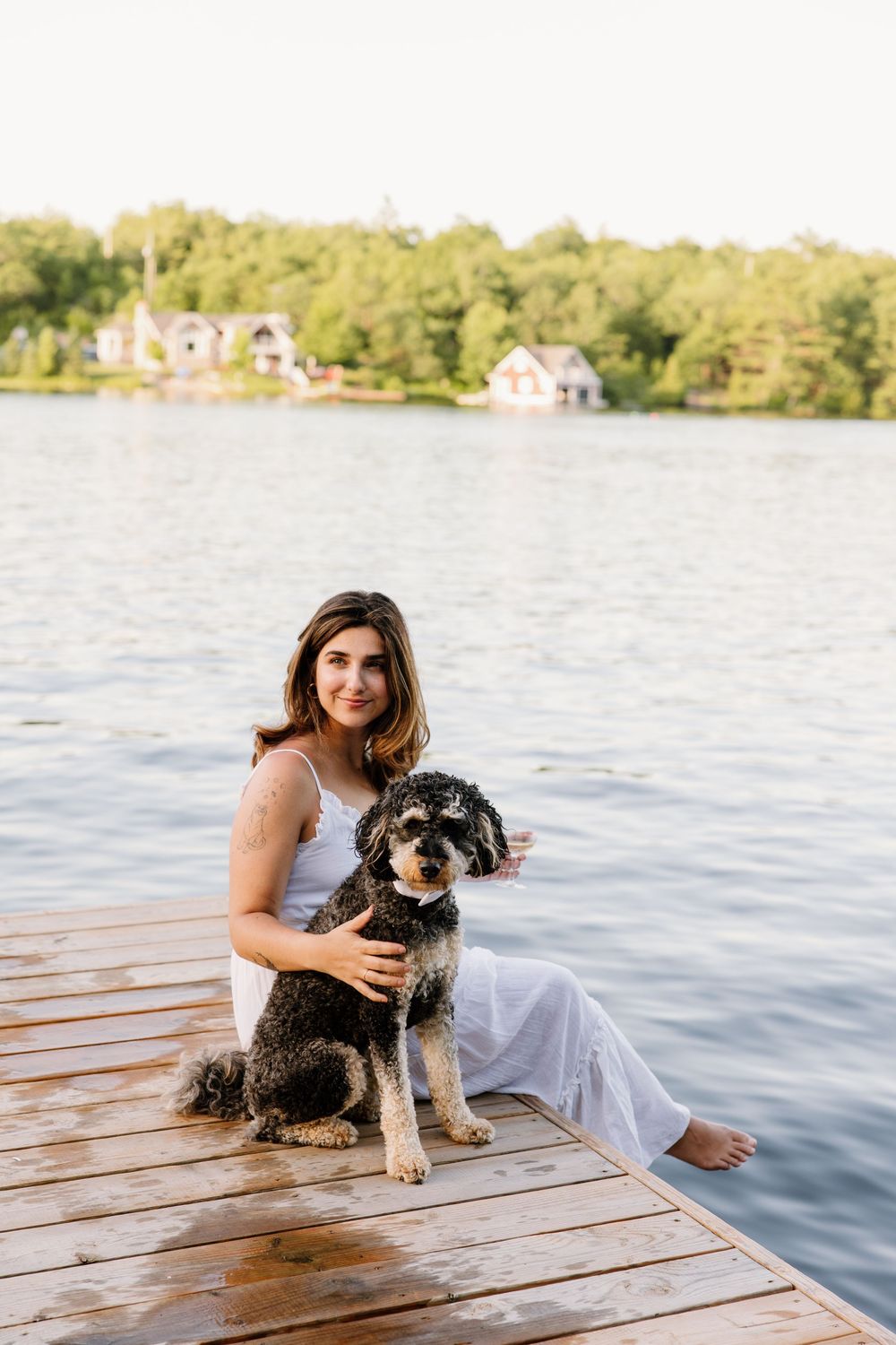 A person in white sits with their black and white dog on a wooden dock by the lake.