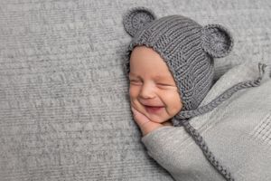 Baby wearing knitted grey hat with ear details smiling while lying on grey fabric.