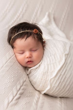 Sleeping newborn baby swaddled in white knit blanket photographed in soft light during studio session.