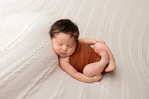 A sleeping newborn baby in a brown knit wrap lies peacefully on soft white fabric during a professional photo session.