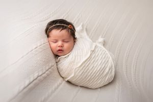 A sleeping newborn baby swaddled in white fabric lying on a soft white surface.