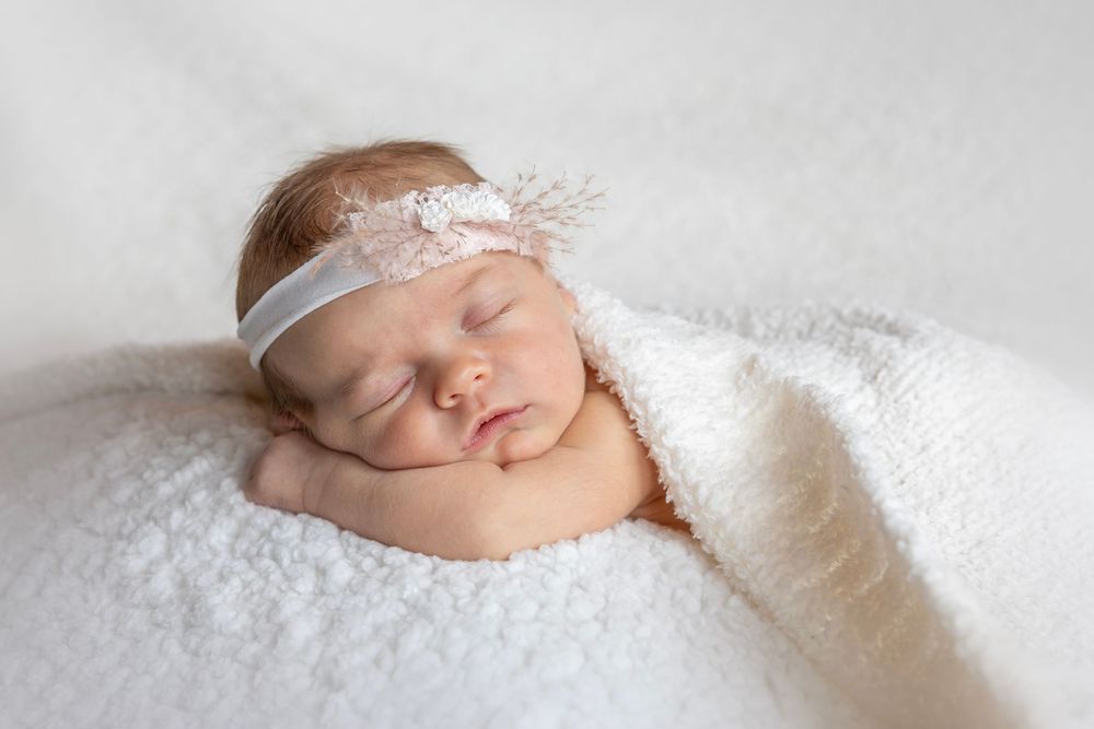 A peaceful sleeping baby lies on soft white fabric wearing a delicate lace headband.