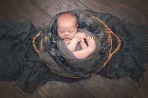 Newborn baby sleeping in round woven basket with dark textured backdrop.