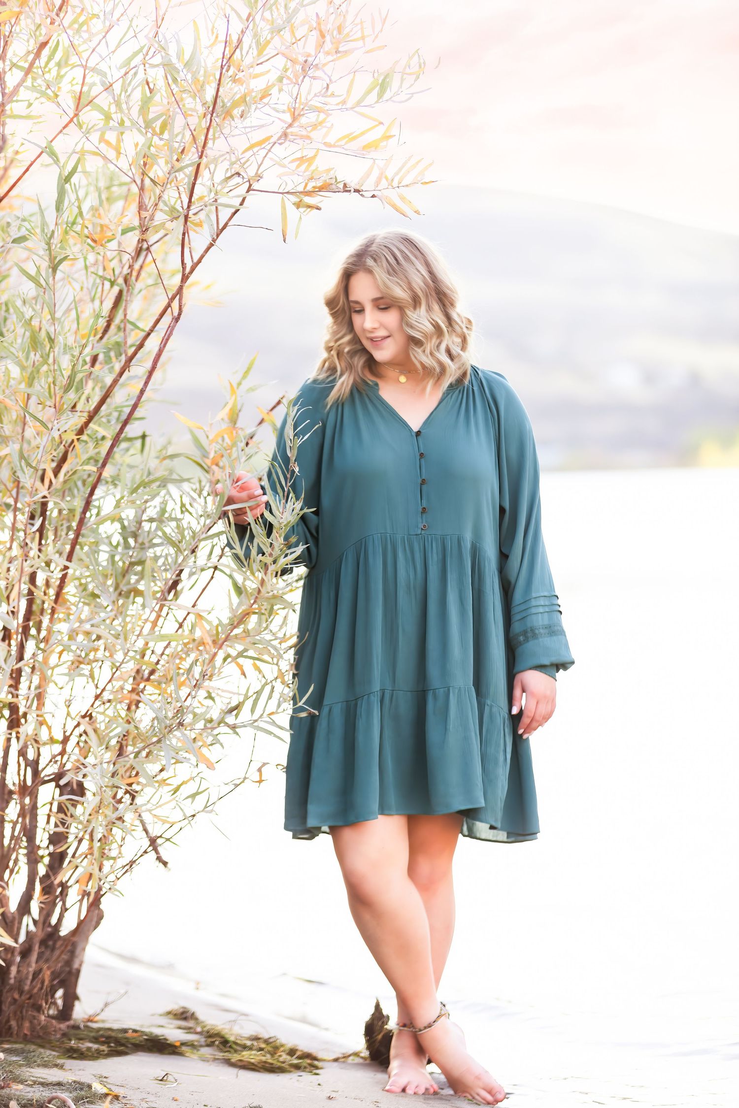 Woman in teal flowy dress poses gracefully by natural foliage.