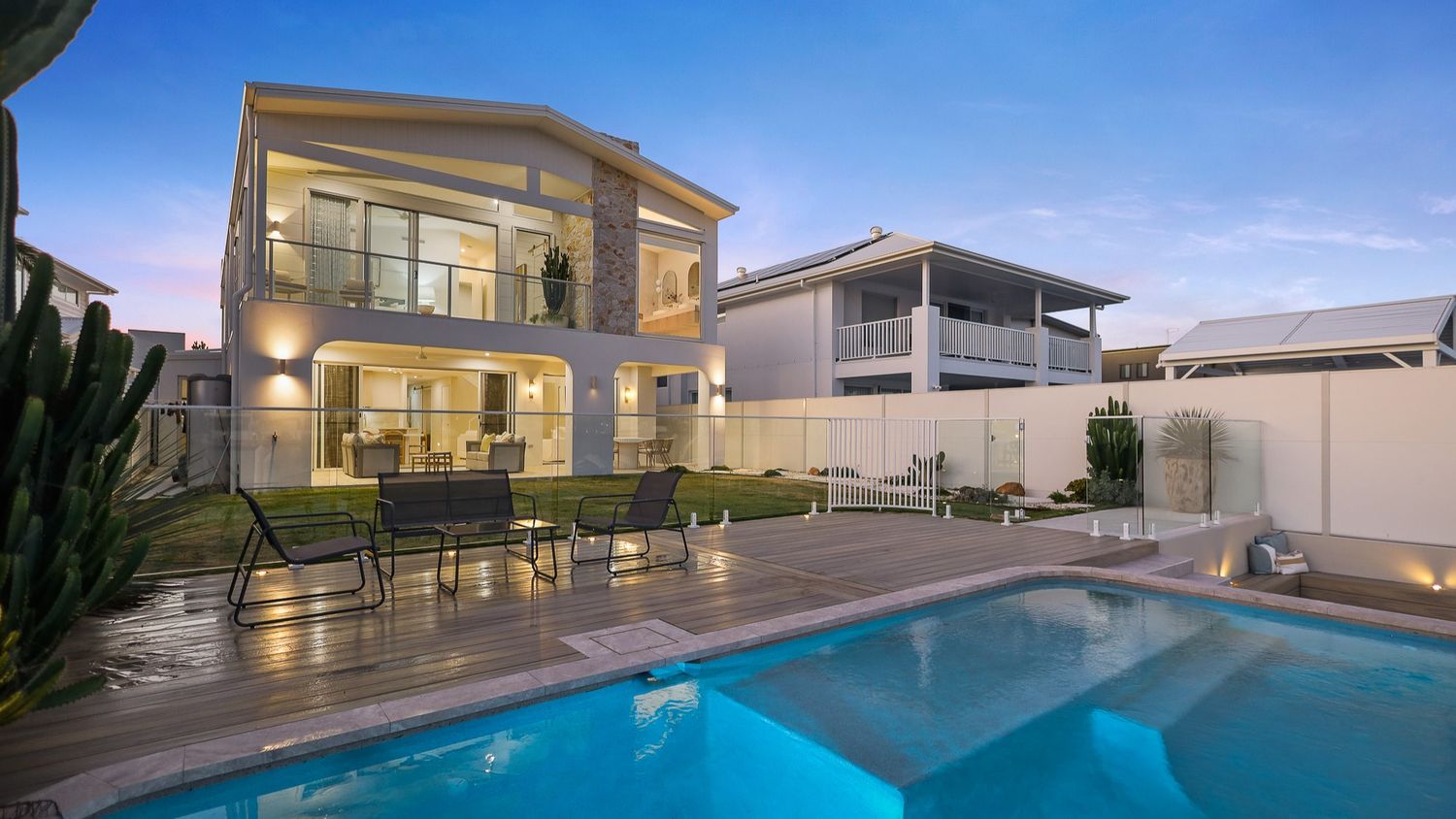 Modern two-story luxury home with illuminated balcony and swimming pool at dusk against blue sky.