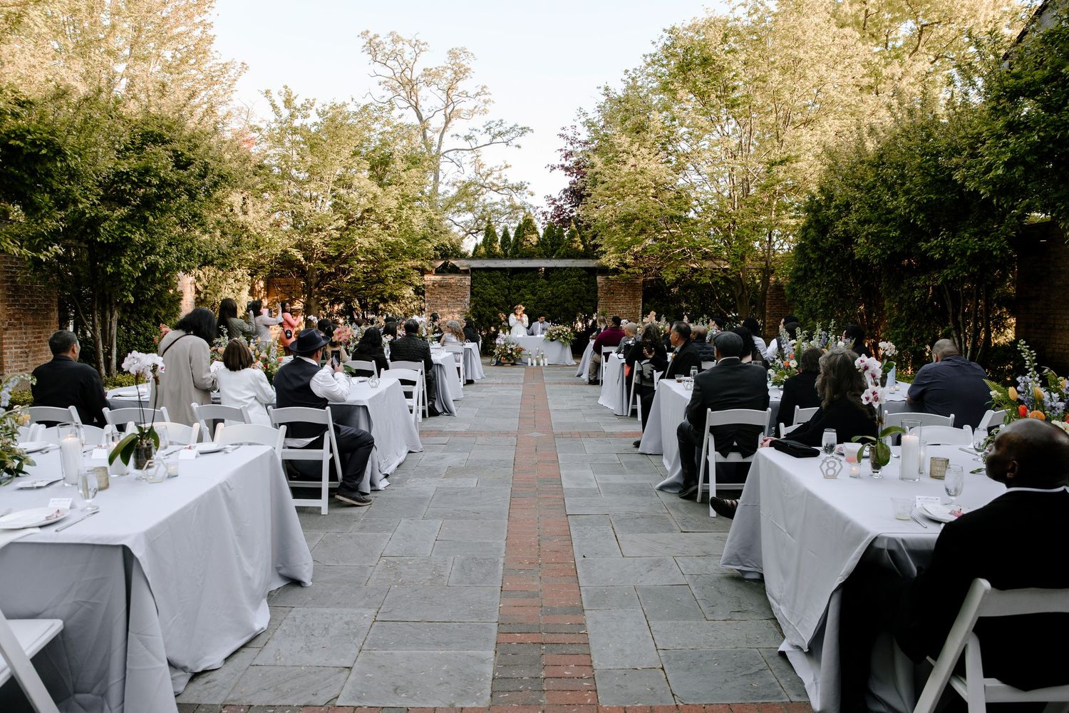 Long outdoor wedding reception tables with white linens arranged under tall trees at dusk.