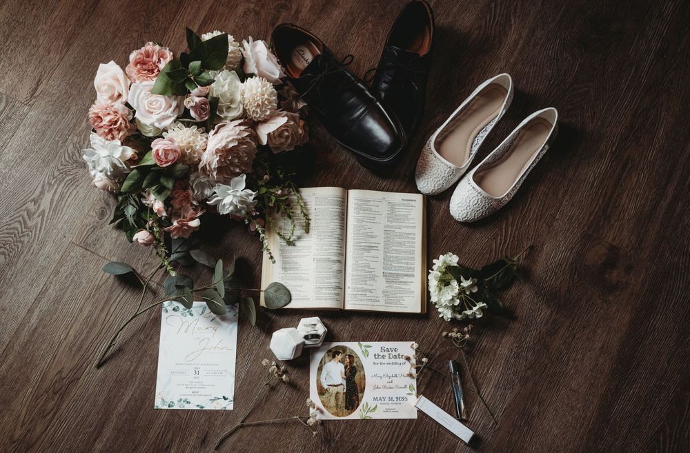 Wedding details flatlay with flowers, invitation, and rings on dark wooden surface.