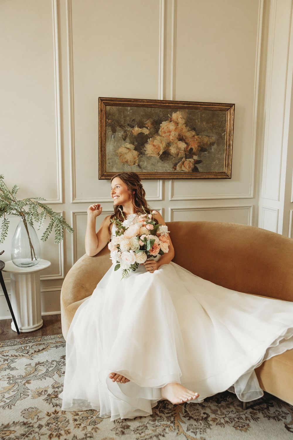 Bride in flowing white gown relaxing on brown couch in elegant room with wall paneling.