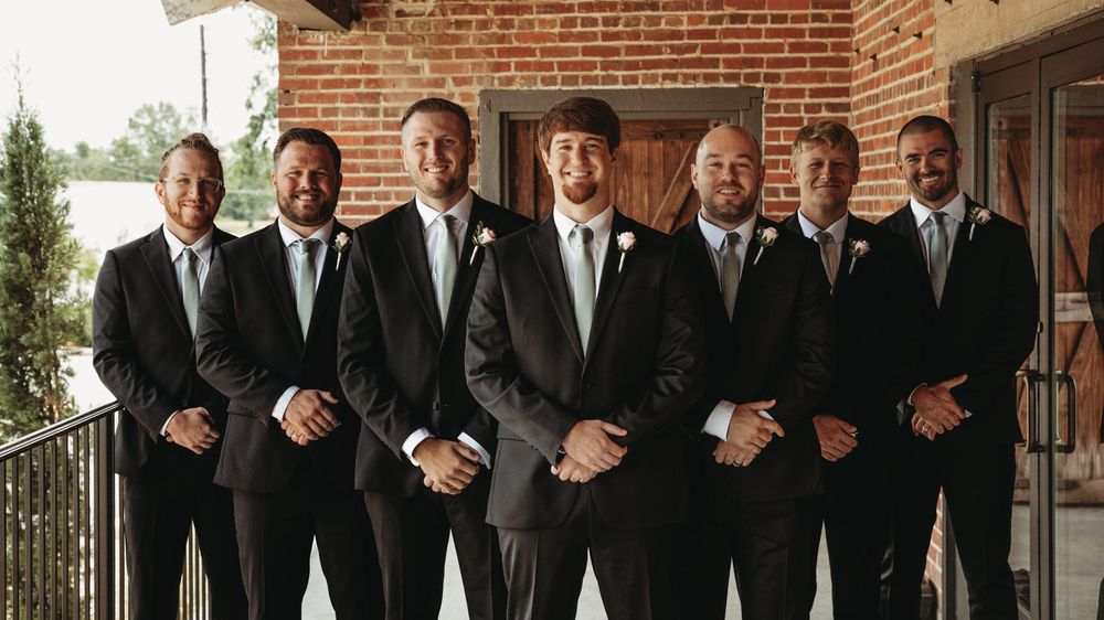 Groomsmen in black tuxedos line up for a formal wedding photo against a brick wall.