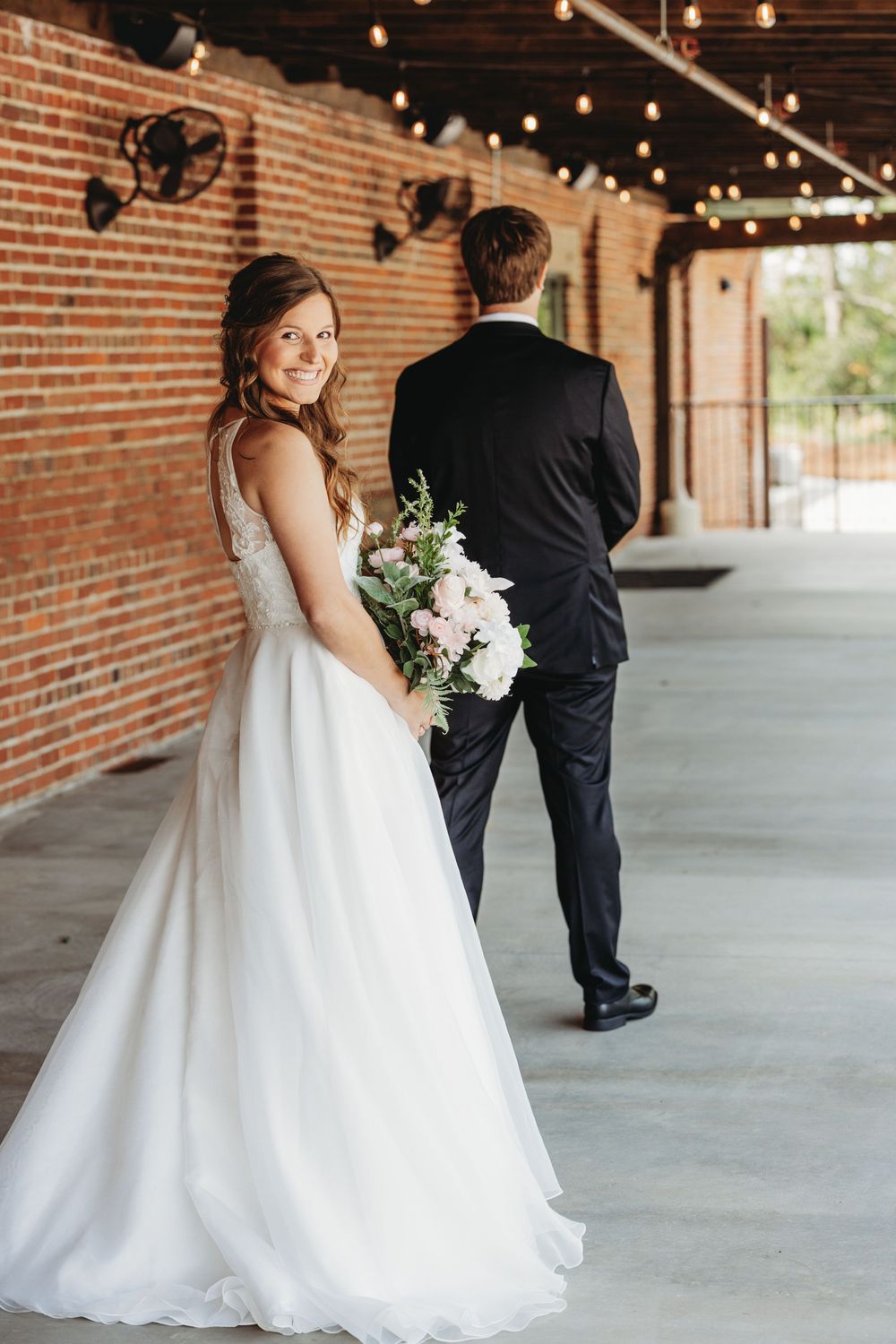 First look wedding moment under string lights in brick corridor.