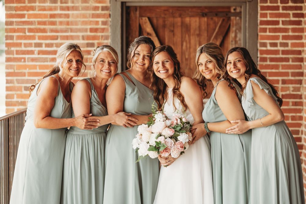 Bridesmaids in sage green dresses pose together in a line at a rustic brick venue.