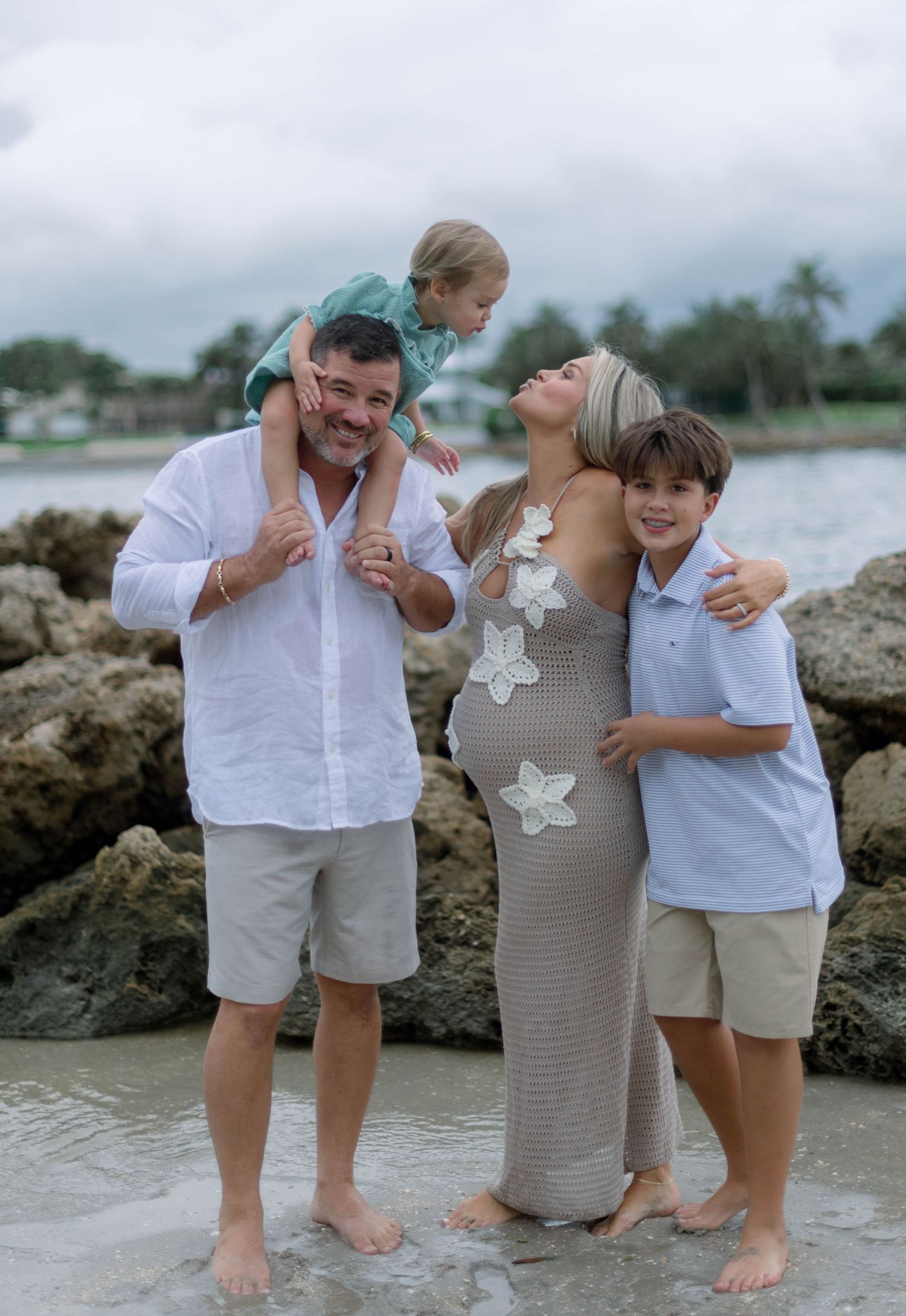 A family of four poses together barefoot on rocky beach terrain with palm trees and water in the background.