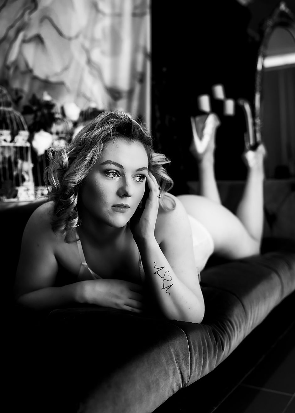 A dramatic black and white boudoir photograph of a model reclining on dark fabric in an artistic pose.