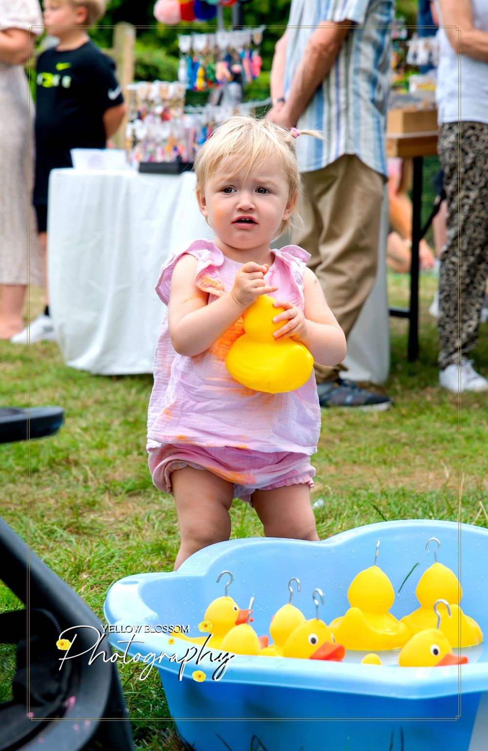 A small child plays with yellow rubber ducks in a blue wading pool on grass during a sunny outdoor gathering.