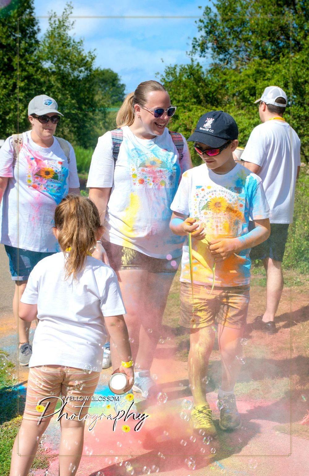 People in white shirts covered in colorful powder paint walk along a sunny outdoor path during a color run event.