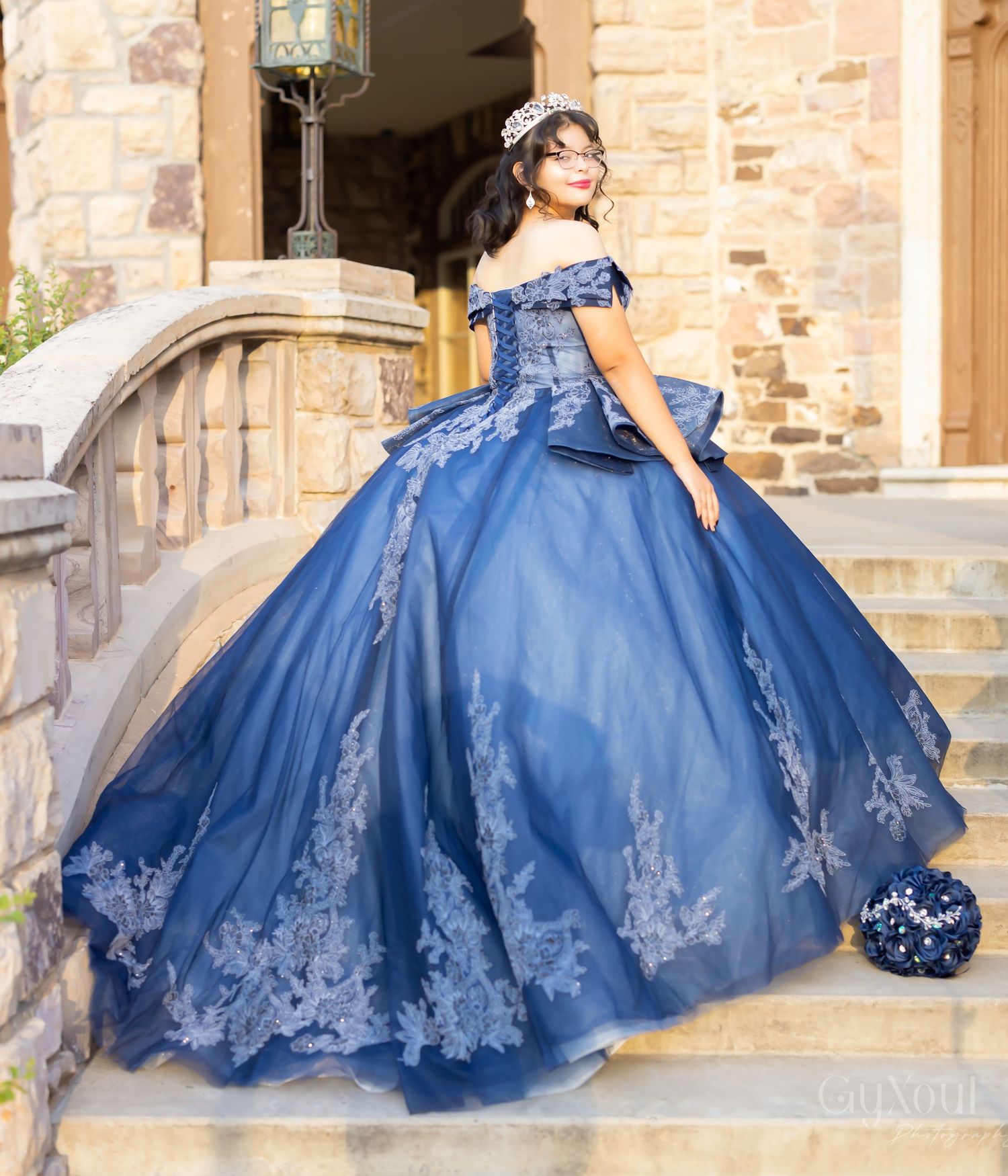 Stunning navy blue quinceanera ball gown with off-shoulder design and intricate silver lace details on stone steps.