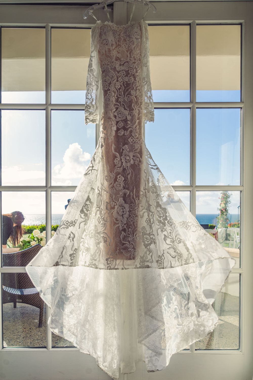 An elegant lace wedding dress hangs in front of a window with a scenic ocean view backdrop.