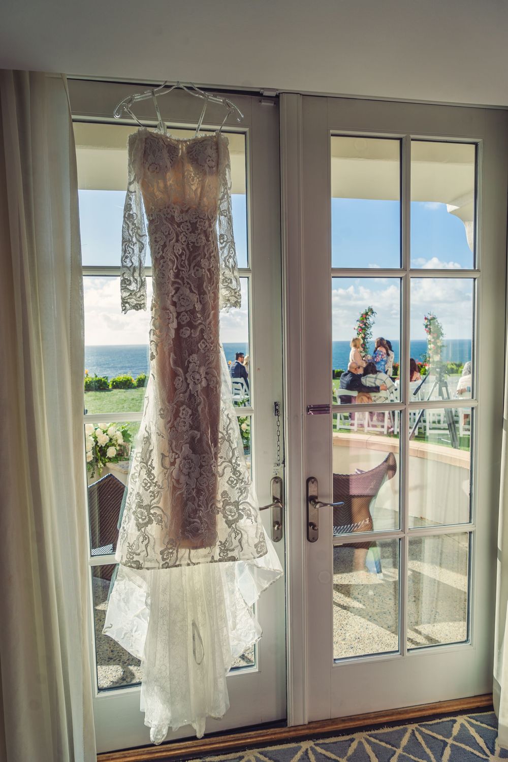 A delicate lace wedding dress hangs in front of French doors with an ocean view on a sunny day.