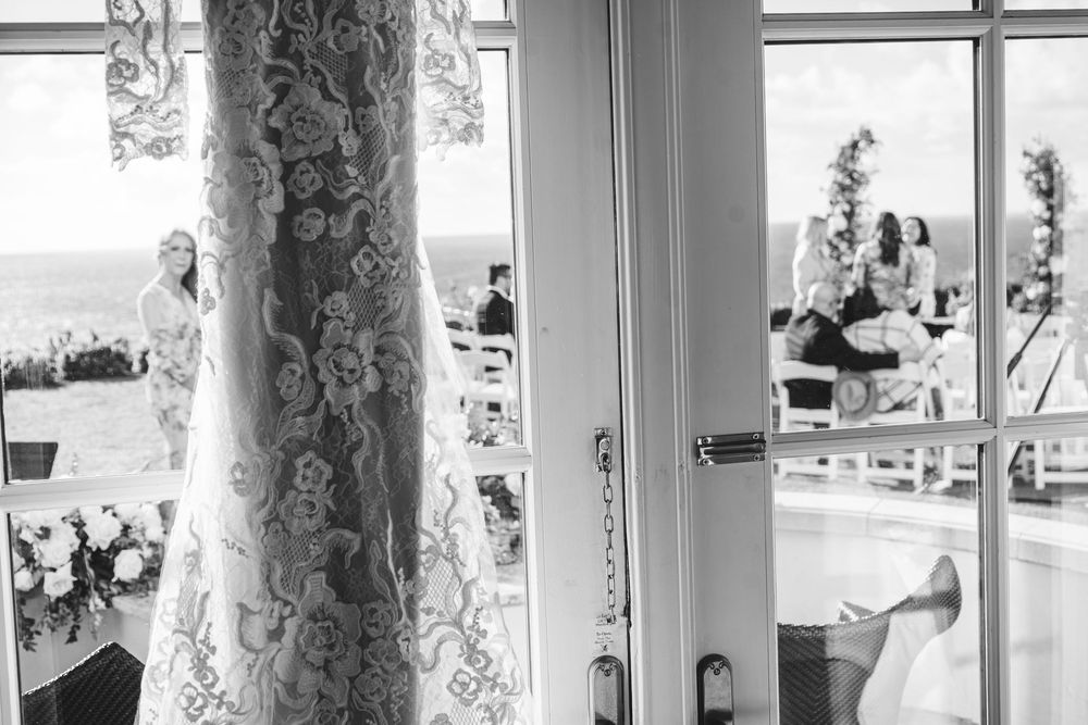 A bride in a white lace wedding dress stands by sunlit windows with flowing sheer curtains.