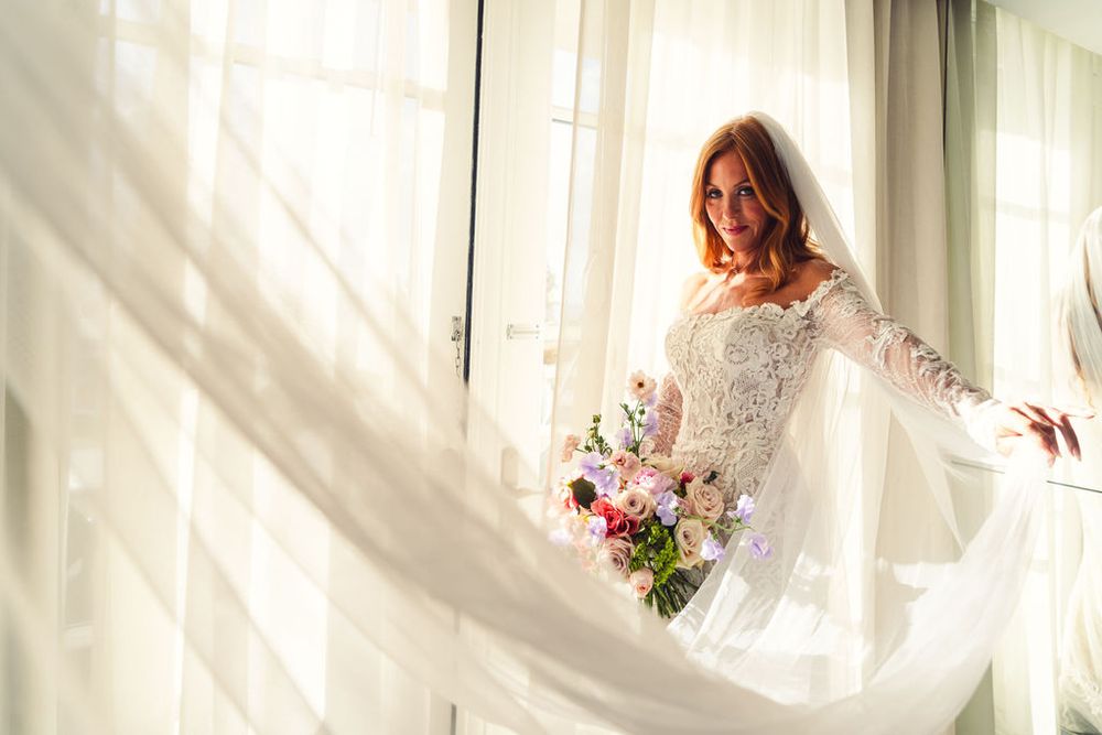 A bride in long sleeve lace wedding dress holds a bouquet by sunlit window with flowing veil.