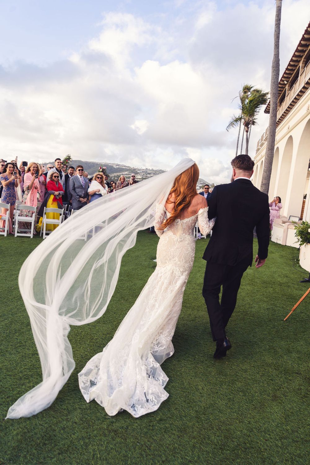 Long flowing wedding veil billows dramatically in the wind as couple walks on grass lawn during outdoor ceremony.