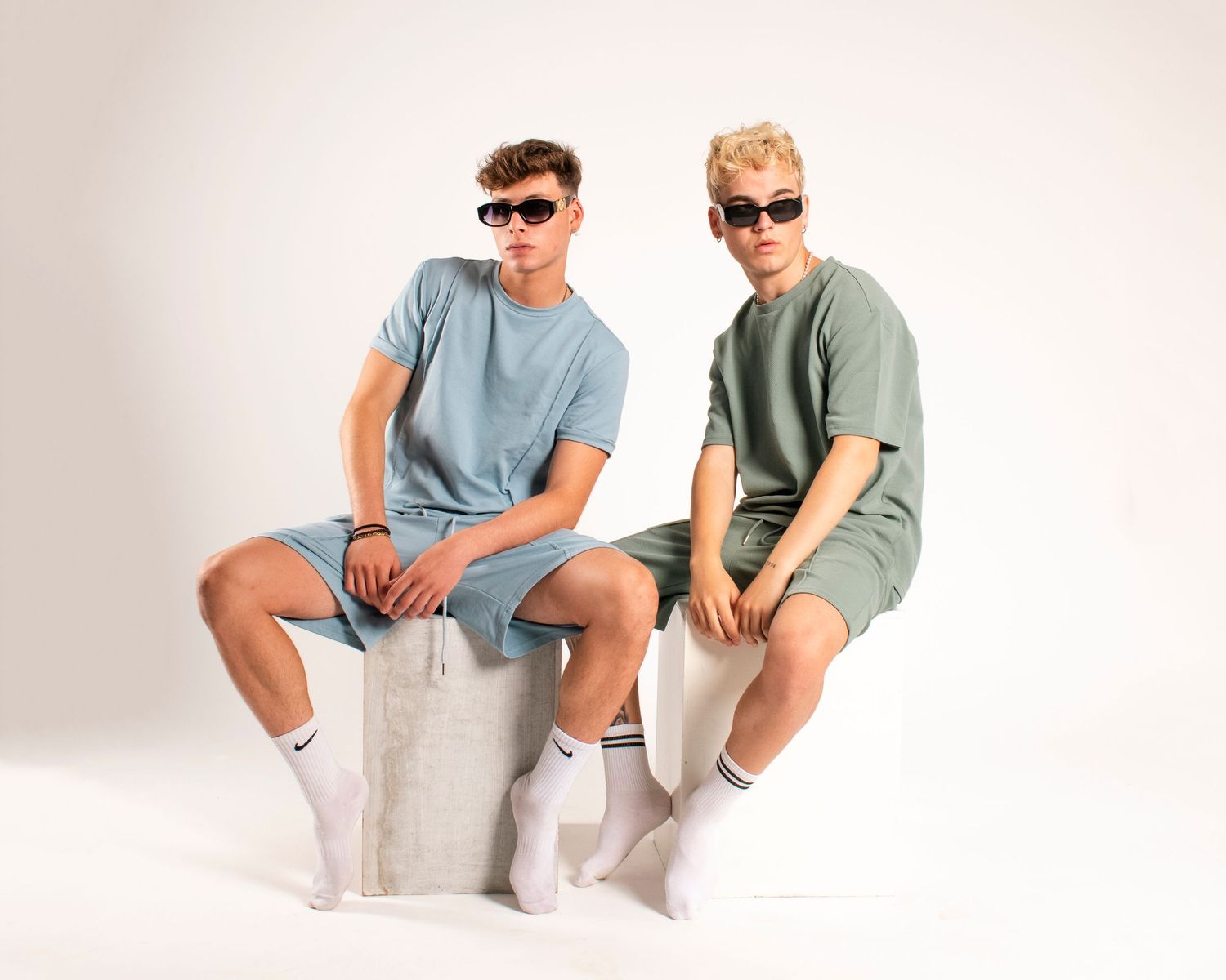 Two models in monochromatic outfits and sunglasses seated on white platform.