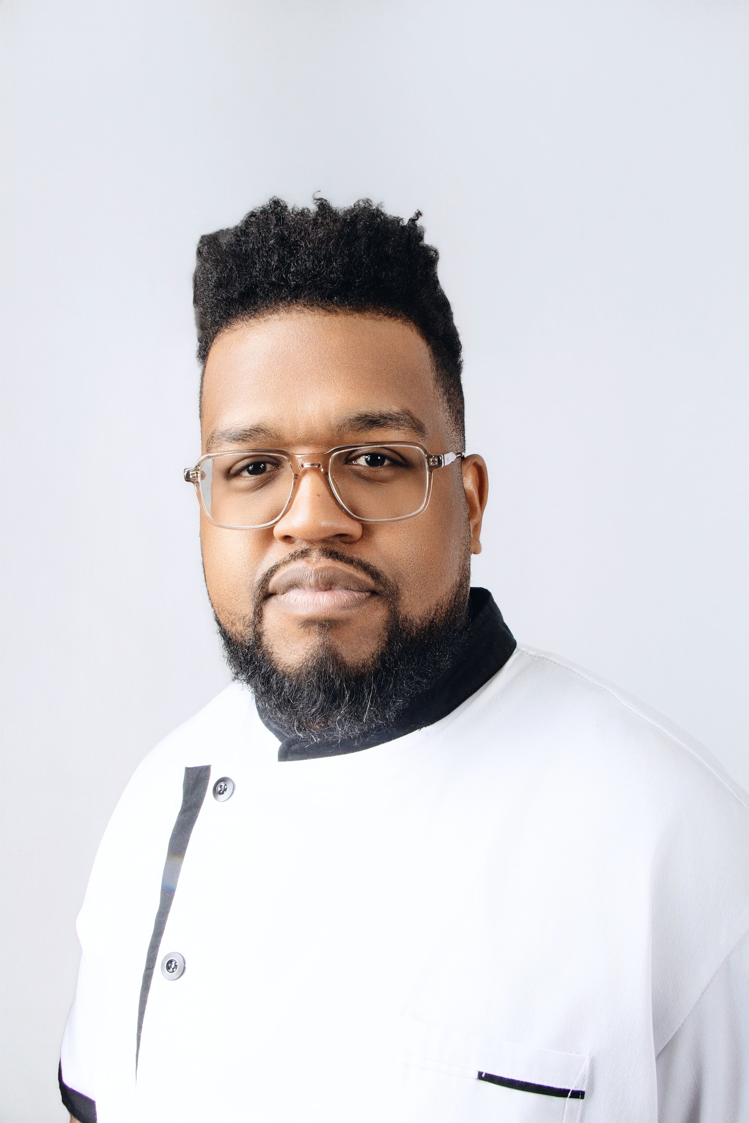 Professional headshot in white chef coat with black trim and glasses against light background.