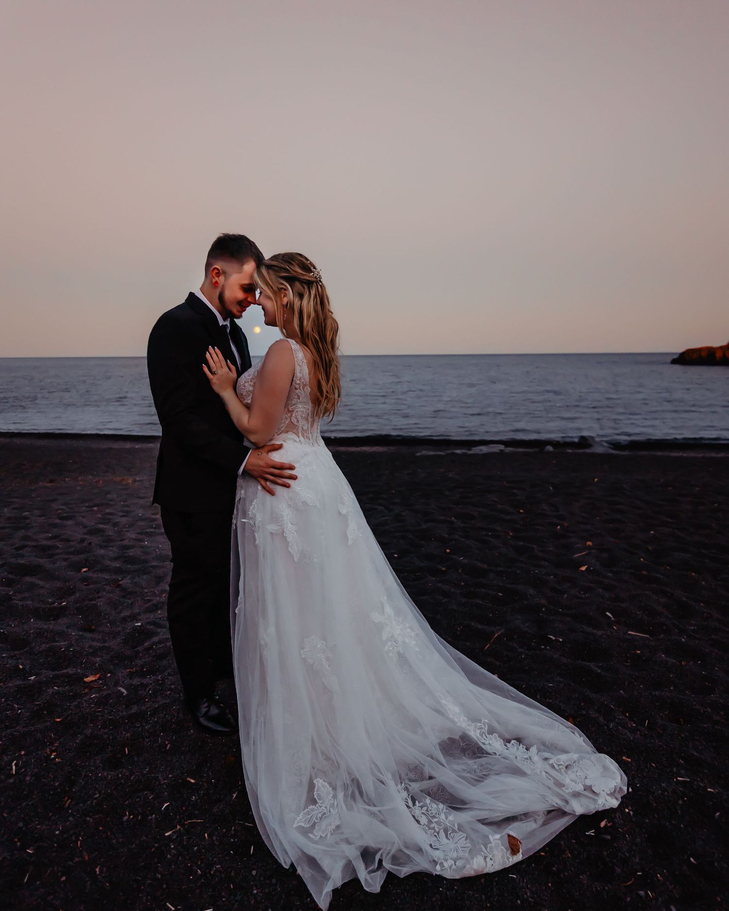 A romantic sunset beach wedding photo with a stunning lace dress train on dark sand.