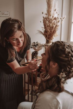 Makeup artist applies cosmetics to a client in a warmly lit room with dried pampas grass decor in the background.