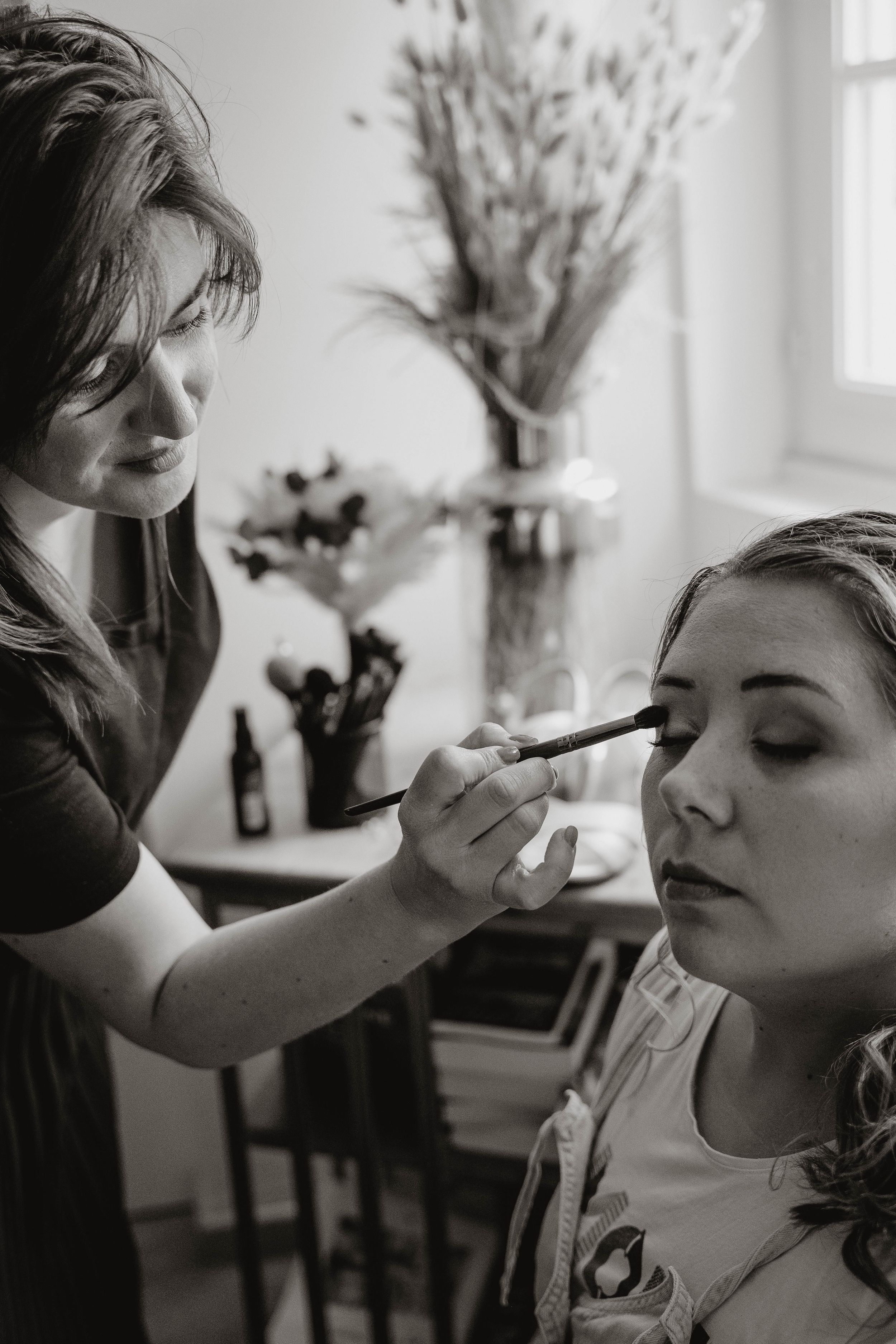 Black and white photo series showing makeup being applied to a model's face in a bright interior setting with flowers.