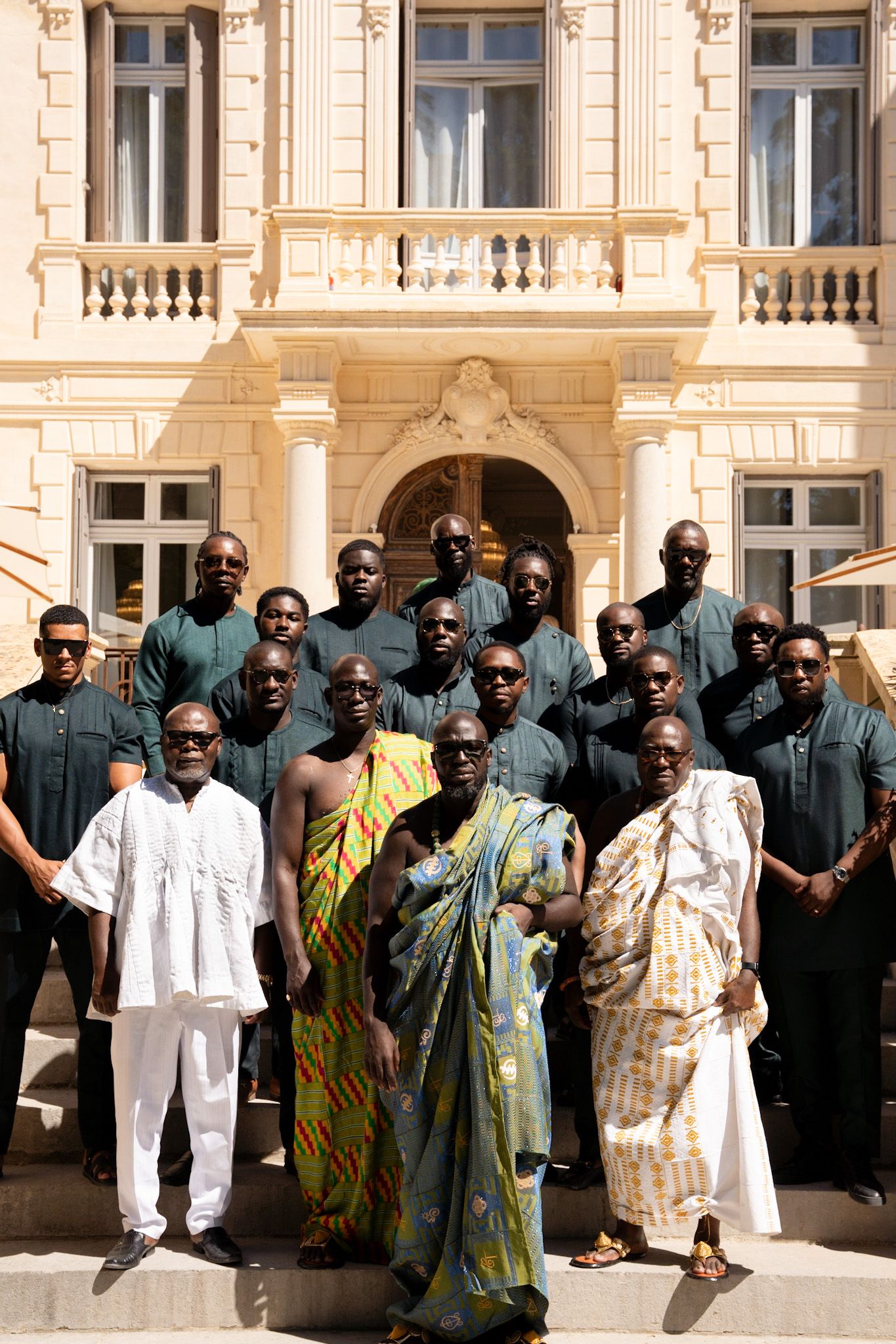 A group of people wearing traditional Ghanian kente cloth and formal attire pose together outside an ornate building.