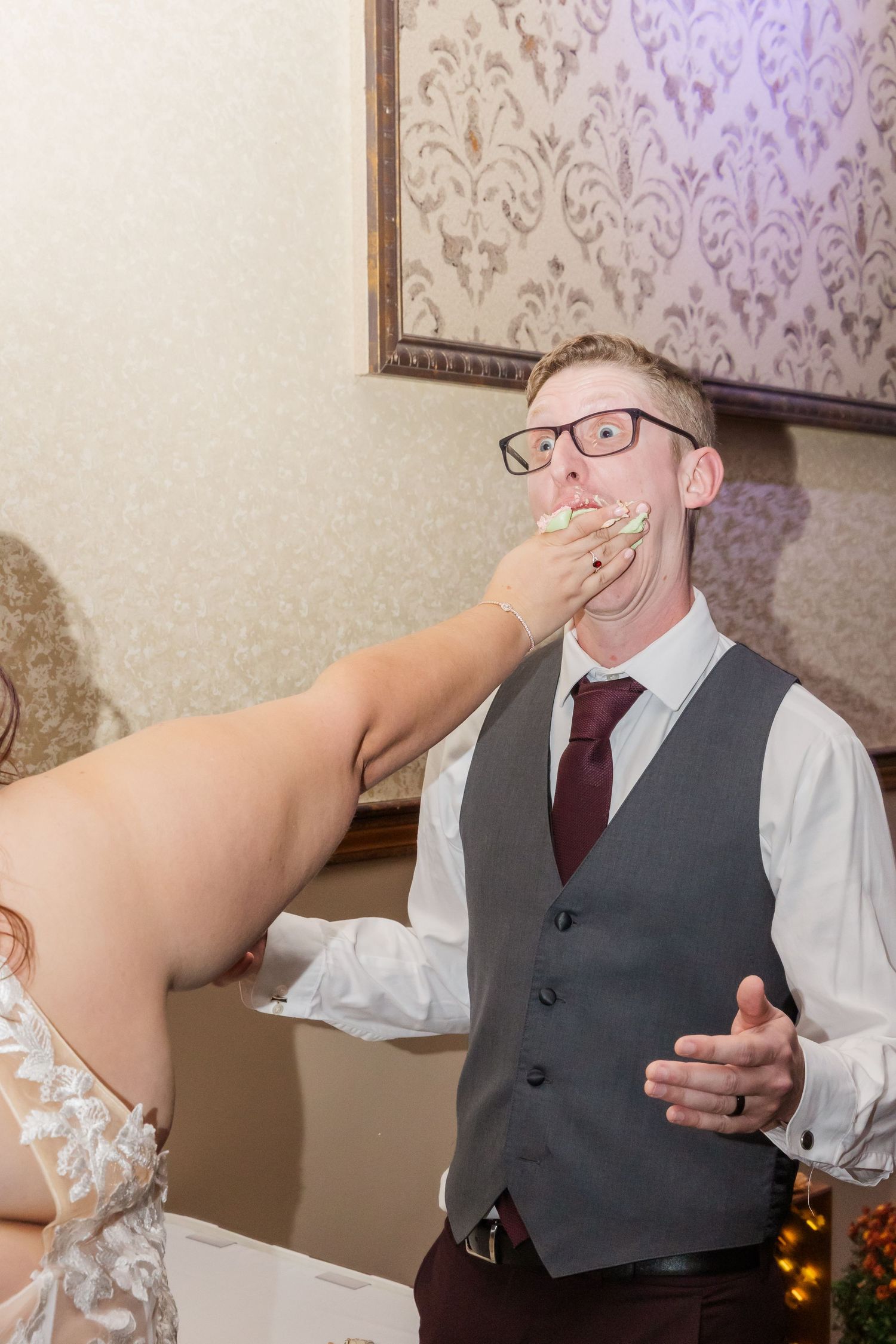 Someone feeds wedding cake to a person wearing a grey vest and burgundy tie at a reception.