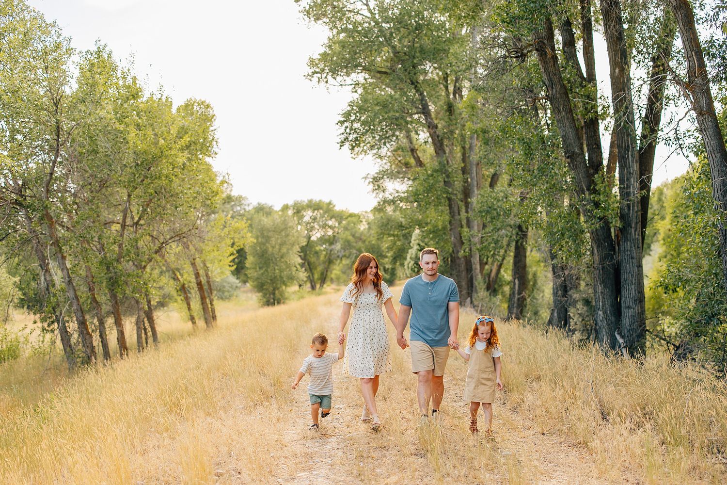 Family of four walking hand-in-hand on a sunny dirt path surrounded by tall green trees and golden grass.