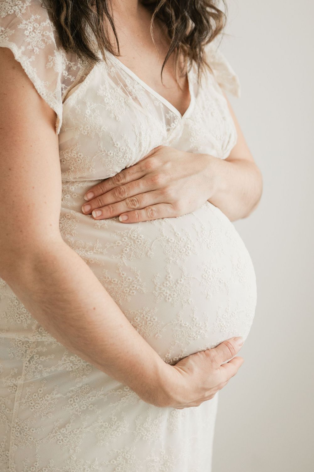 Pregnant woman in white lace dress cradling her baby bump with both hands against a light background. Cornwall Maternity Photographer