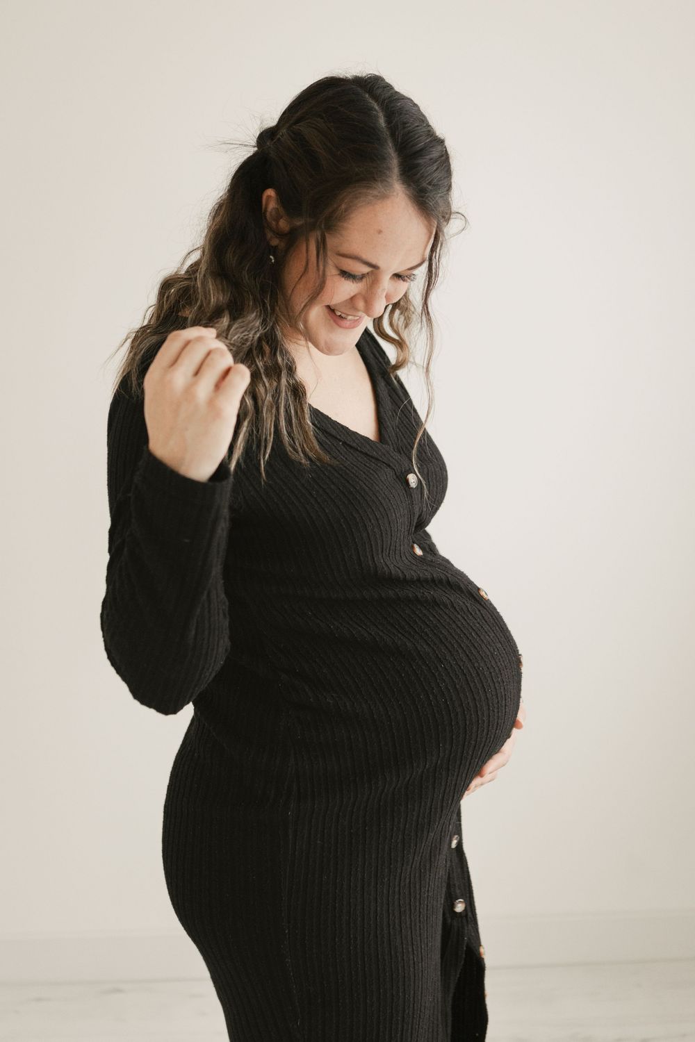 Pregnant woman in black ribbed dress smiles down at her baby bump against a white studio background. Cornwall Maternity Photographer