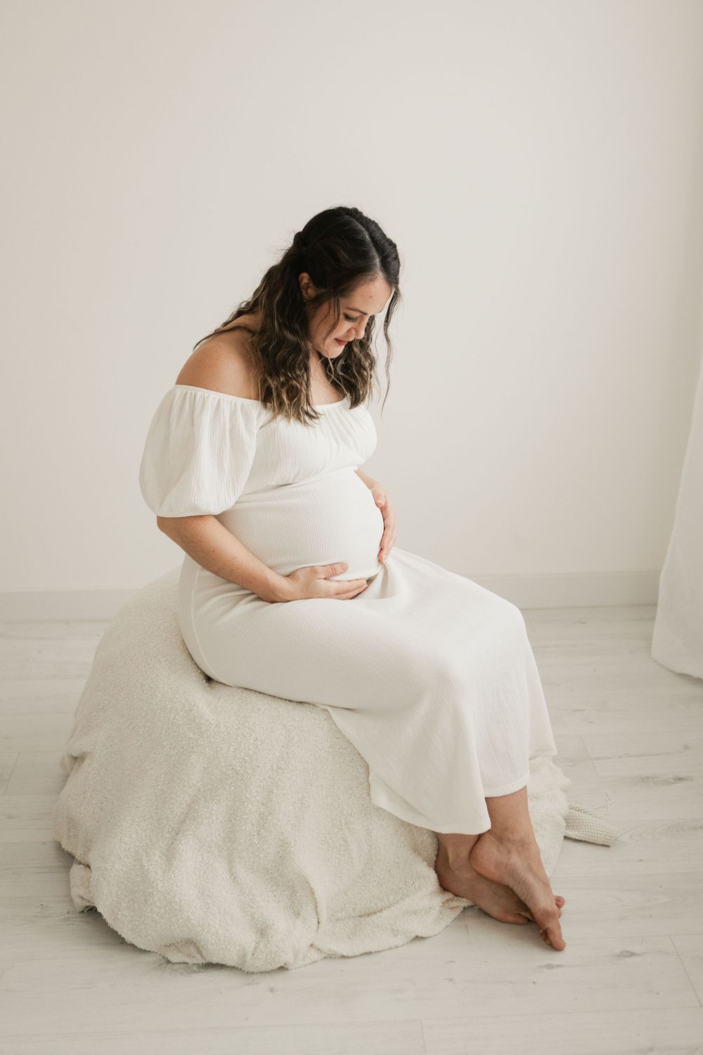 Pregnant woman in white off-shoulder dress sits barefoot, cradling her baby bump in a bright, minimalist studio. Cornwall Maternity Photogra