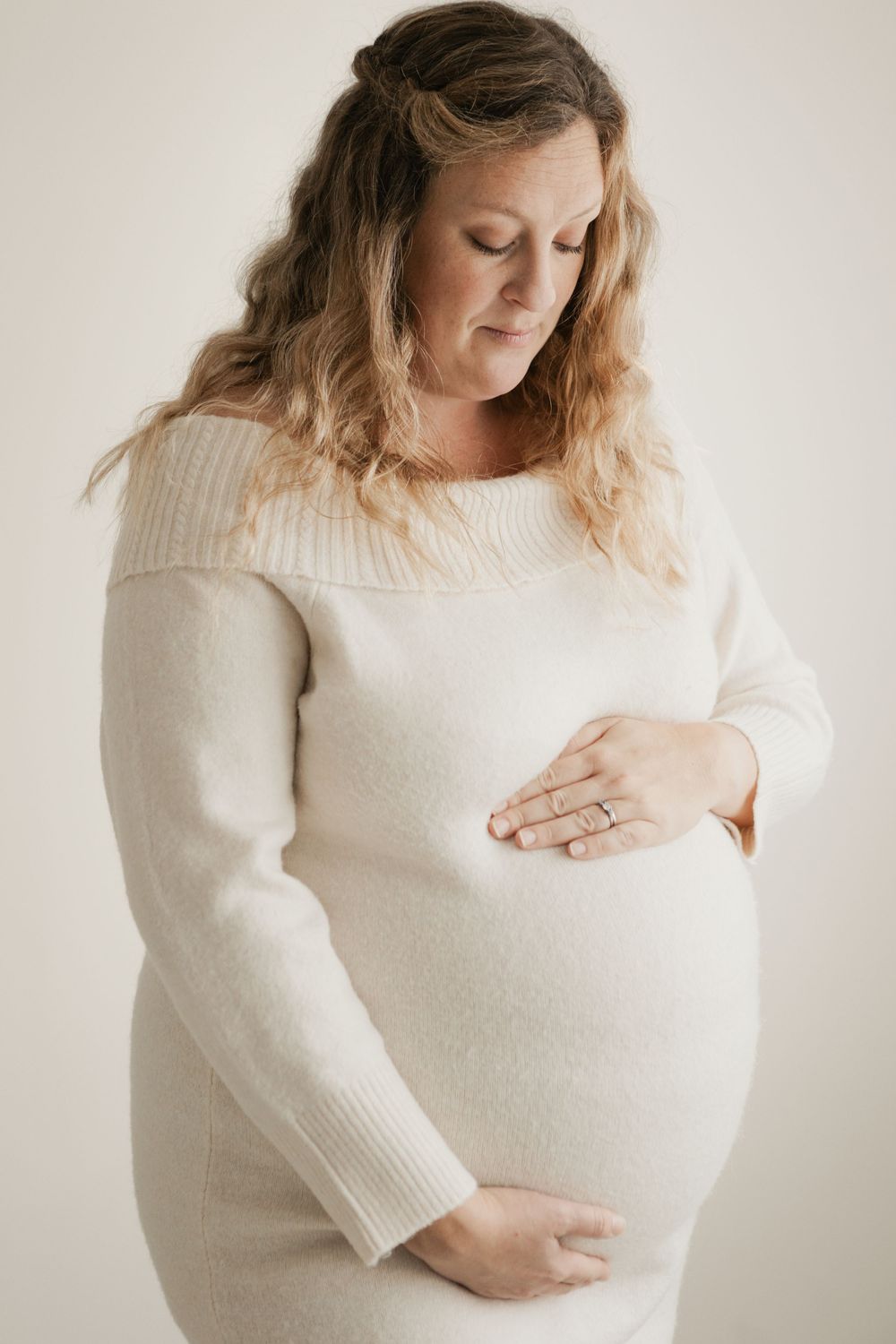Pregnant woman in cream off-shoulder sweater dress cradling baby bump, studio maternity portrait. Cornwall Maternity Photographer