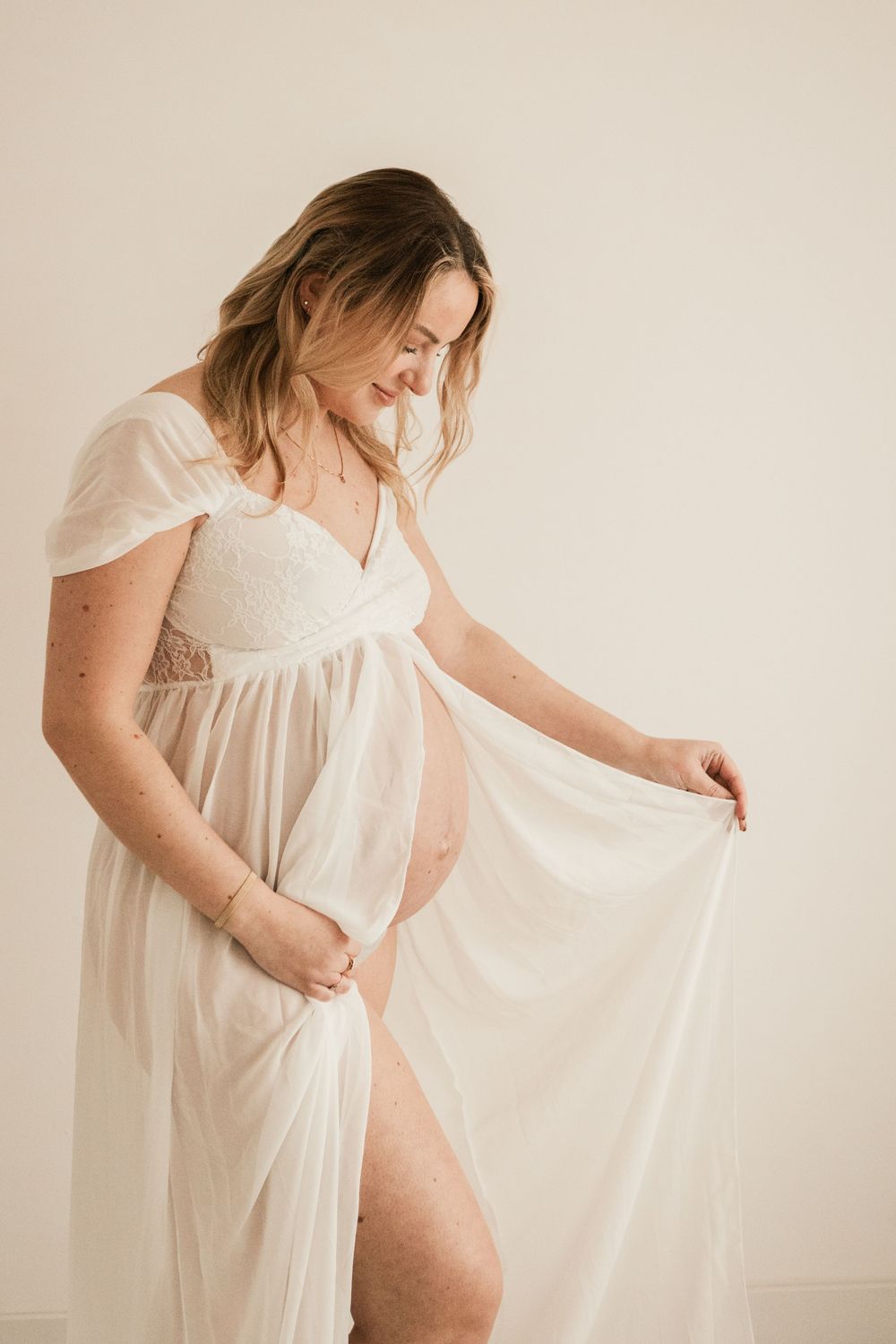 Pregnant woman in flowing white maternity gown holding fabric open, studio portrait photography. Cornwall Maternity Photographer