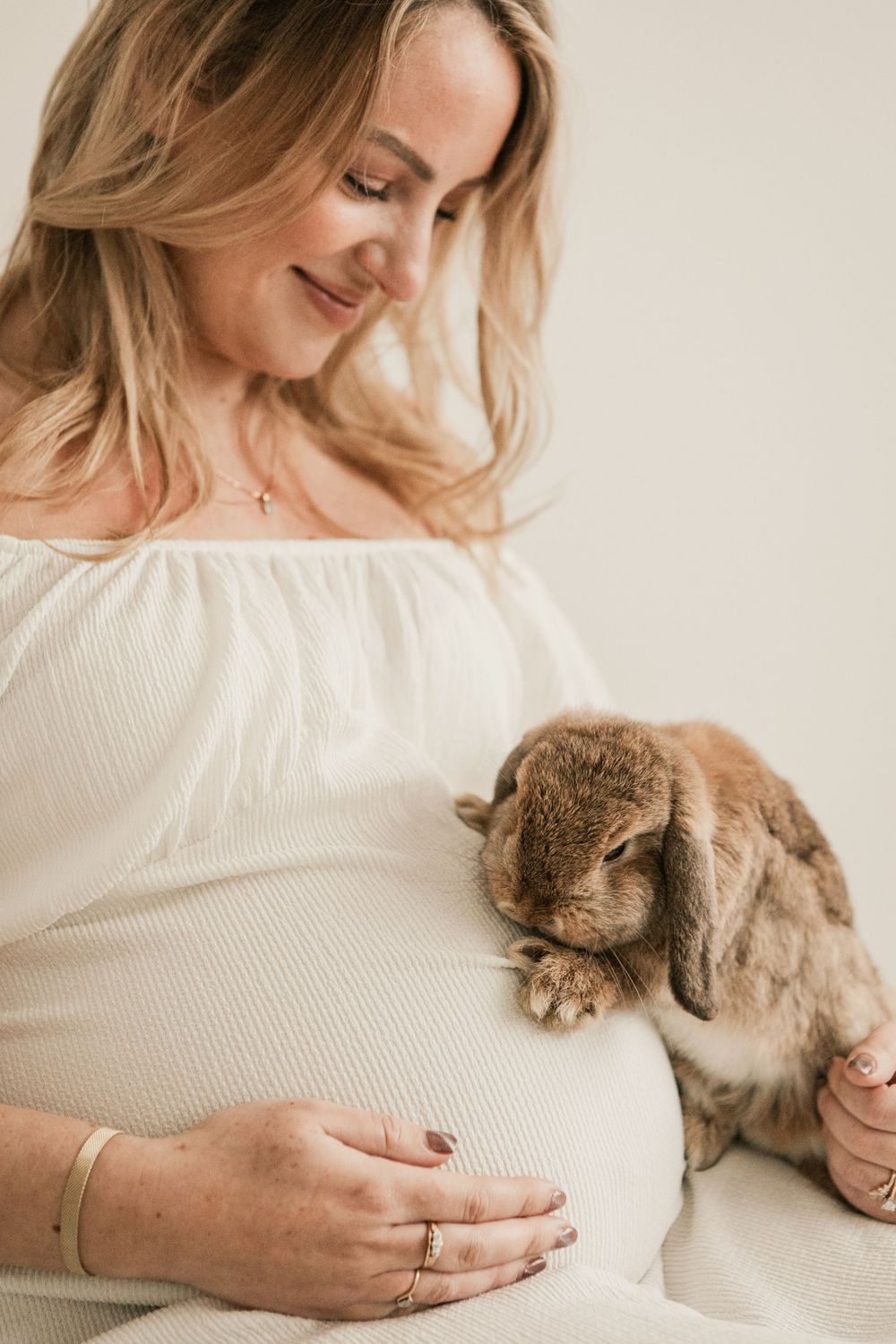 Smiling pregnant woman in white dress holding a brown lop-eared rabbit against her baby bump in Cornwall maternity studio