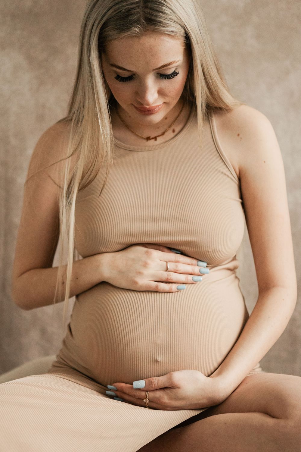 Pregnant woman in beige dress cradling her baby bump, sitting against neutral background in South East Cornwall maternity photography studio